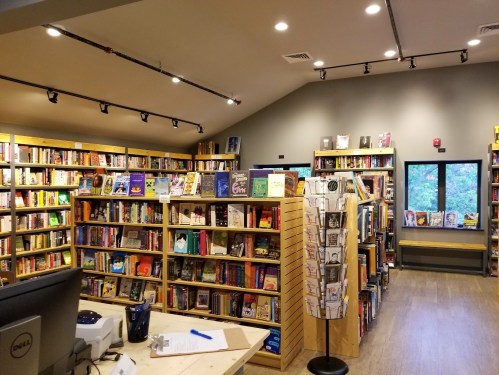 A cozy bookstore interior with wooden shelves filled with books, a checkout counter, and a reading area by the window.
