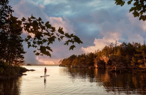 A person paddleboarding on a calm lake at sunset, surrounded by trees and rocky cliffs.