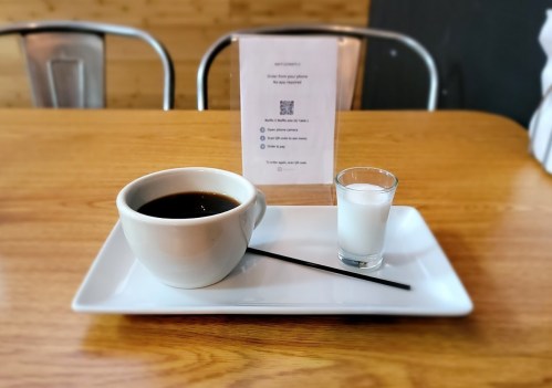 A white bowl of coffee with a black straw, accompanied by a small glass of milk, on a wooden table.