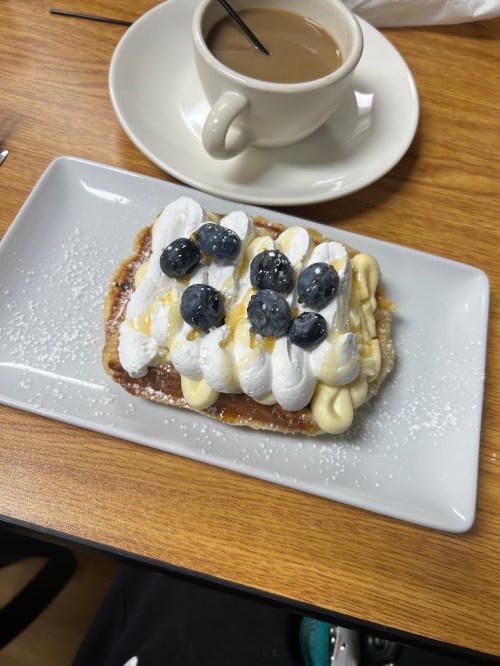 A dessert on a plate topped with whipped cream, blueberries, and a dusting of powdered sugar, alongside a cup of coffee.