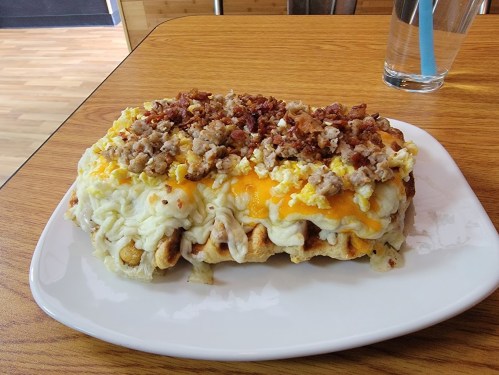 A large dish featuring a waffle topped with scrambled eggs, cheese, and crumbled bacon on a wooden table.