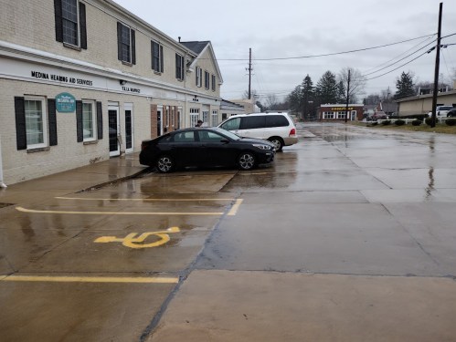 A wet parking lot with a black car and a white van, alongside a building labeled "Media Healing and Services."
