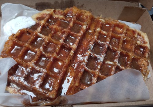 A partially eaten waffle drizzled with syrup, served in a takeout box with a dollop of whipped cream.