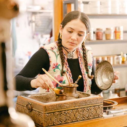 A young woman with braids carefully prepares food in a rustic kitchen, using traditional utensils and a decorative box.