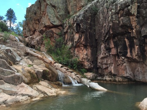 A serene waterfall cascades over rocks into a calm pool, surrounded by rugged cliffs and greenery.
