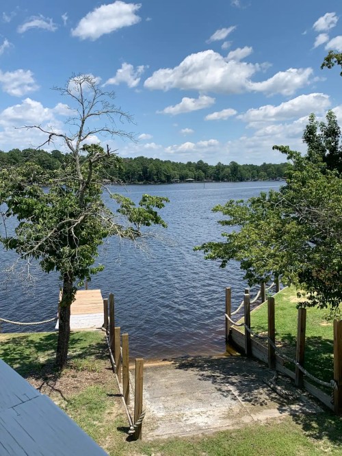 A serene lake view with trees, a wooden dock, and a clear blue sky dotted with clouds.
