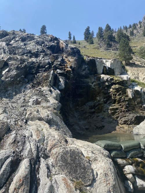 A rocky landscape with a small waterfall cascading into a pool, surrounded by trees and blue sky.