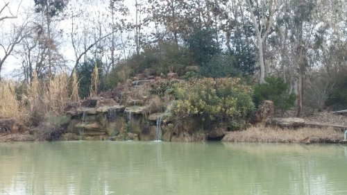 A serene pond with a rocky waterfall and lush greenery surrounding it, set against a cloudy sky.