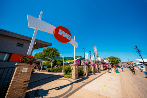 A sunny outdoor fair scene with a large sign reading "We" and colorful flower arrangements along the walkway.