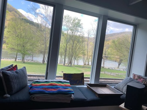 A cozy window seat with colorful blankets overlooks a river and trees, creating a serene outdoor view.