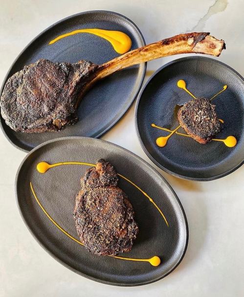 A beautifully presented meal featuring a large bone-in steak and smaller cuts, garnished with yellow sauce on black plates.