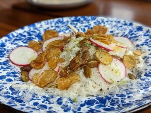 A colorful plate of salad featuring thin noodles, radish slices, crispy toppings, and seeds on a decorative blue dish.