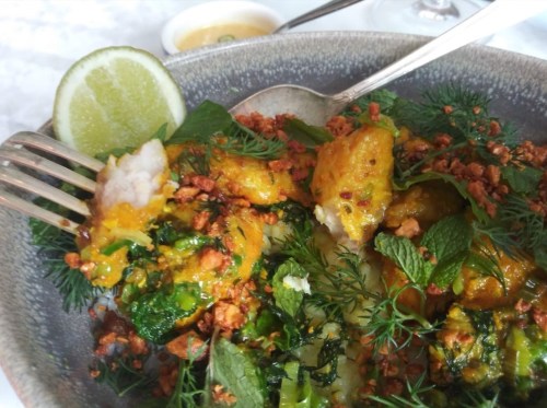 A close-up of a colorful dish featuring fish, herbs, and crispy toppings, garnished with a lime wedge.