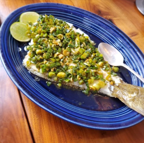 A blue plate holds a whole fish topped with green onions, herbs, and lime slices, garnished with a spoon on the side.