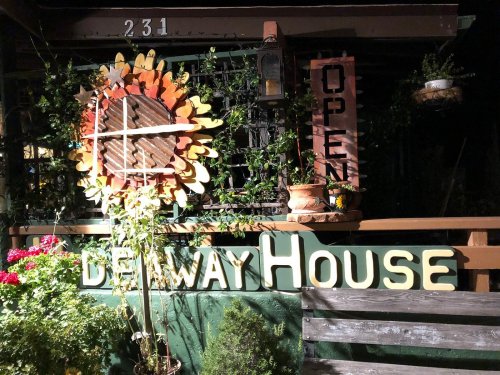 A cozy entrance with a sunflower decoration and a sign reading "Delway House" and "Open," surrounded by plants.