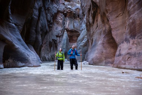 Two hikers wade through a river in a narrow canyon, surrounded by towering rock walls, looking up in awe.