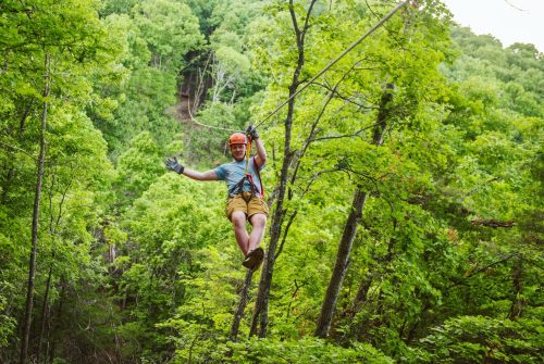 A person in a helmet zip-lining through a lush green forest, surrounded by trees and hills.