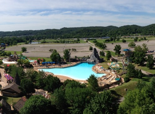 Aerial view of a large water park with a blue pool, slides, and surrounding greenery on a sunny day.