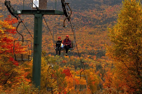 Two people on a ski lift surrounded by vibrant autumn foliage in a mountainous landscape.