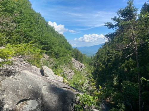 A scenic view of a rocky valley surrounded by lush green trees under a blue sky with scattered clouds.