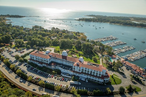 Aerial view of a coastal hotel with a marina, surrounded by trees and water under a clear blue sky.