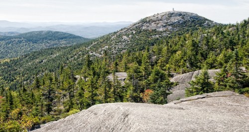 A rocky mountain peak surrounded by dense green forests and distant hills under a clear sky.