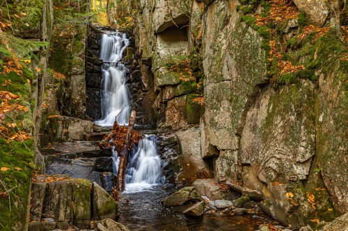 A serene waterfall cascades over rocky cliffs, surrounded by autumn foliage and tranquil water.