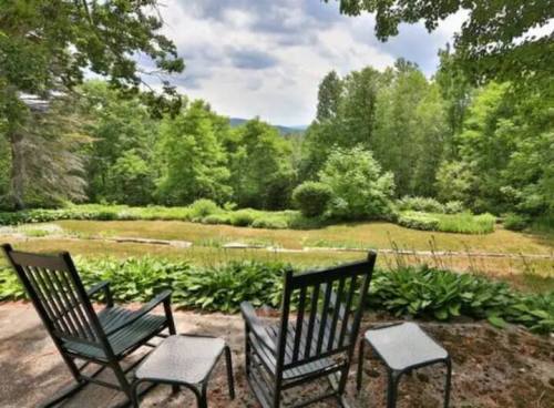 A serene outdoor scene featuring three chairs overlooking a lush green landscape with trees and a pond.