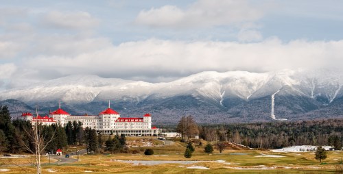 A grand hotel with red roofs sits in front of snow-capped mountains, surrounded by green fields and trees.