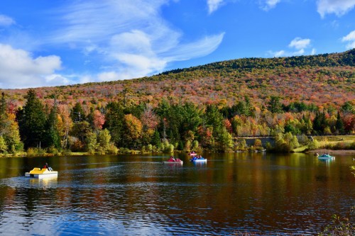 Colorful autumn trees surround a serene lake where people paddle boats under a blue sky with fluffy clouds.