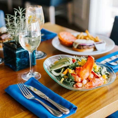 A vibrant salad with lobster, greens, and colorful vegetables, accompanied by a glass of white wine and a burger in the background.