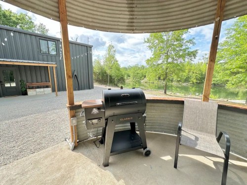 A black grill on a covered patio with a chair, overlooking a gravel path and trees by a pond.