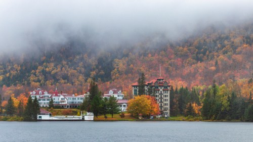 A misty landscape featuring a historic hotel by a lake, surrounded by autumn-colored trees and mountains.