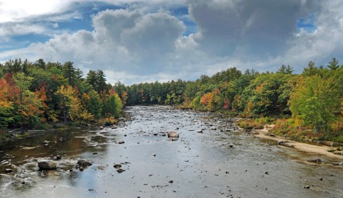 A serene river flows through a forest with vibrant autumn foliage under a cloudy sky.