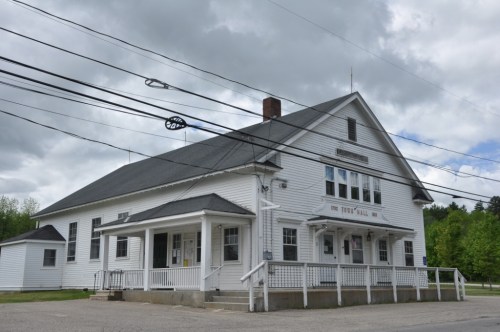 A large white building with a porch, labeled "Town Hall," set against a cloudy sky and surrounded by power lines.