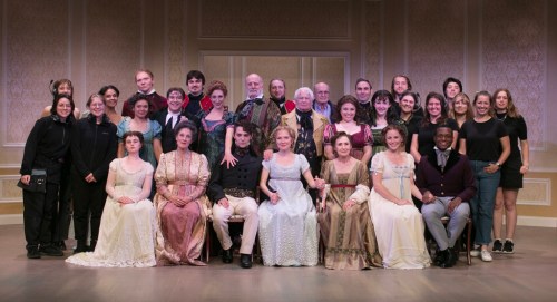 A large group of actors in period costumes poses together on stage, smiling for a photo.