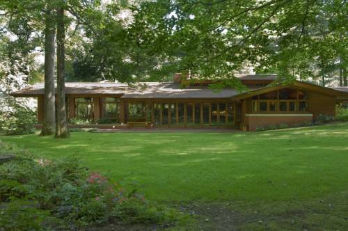 A modern, single-story house with large windows, surrounded by lush greenery and trees.