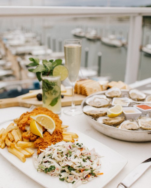 A plate of fried chicken with fries and coleslaw, alongside oysters and drinks, with a marina view in the background.
