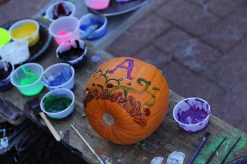 A decorated pumpkin with the letter "A" and colorful paint cups in the background on a wooden table.
