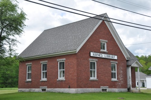 A red brick building with a sloped roof, featuring a sign that reads "Haynes Library" and surrounded by green grass.