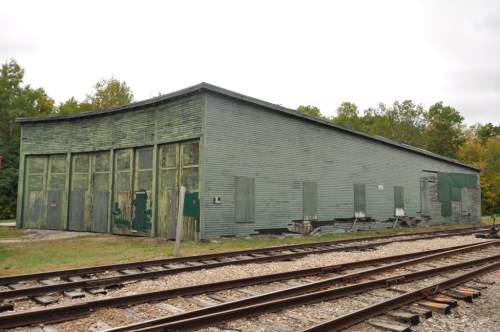A weathered green building with boarded windows, situated near railroad tracks and surrounded by trees.