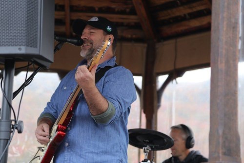 A musician smiles while playing guitar and singing on stage, with a drummer in the background.