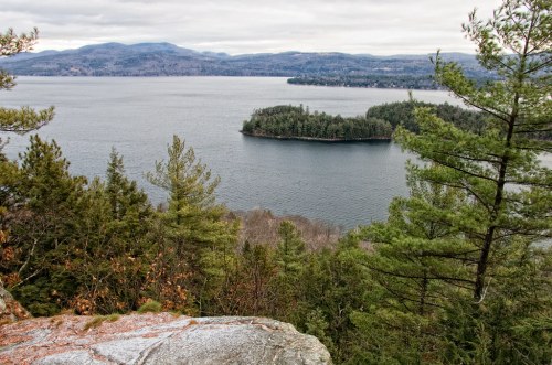 A serene view of a lake surrounded by forested hills and a small island, under a cloudy sky.