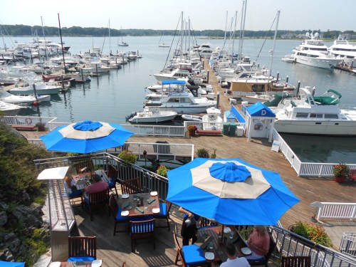 A marina with numerous boats docked, featuring blue umbrellas over outdoor dining areas.