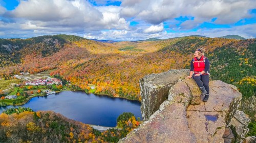 A hiker sits on a rocky ledge overlooking a vibrant autumn landscape with mountains and a lake below.
