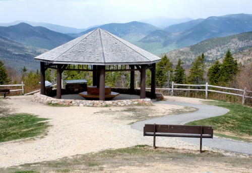 A gazebo with a stone base overlooks rolling green mountains under a cloudy sky, with a path and bench nearby.
