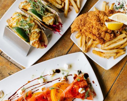 A vibrant spread featuring tacos, fried fish with fries, and a colorful watermelon salad on wooden table.
