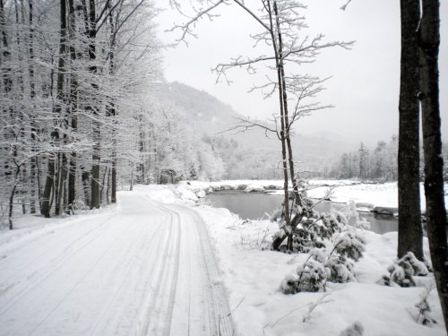 A snowy landscape with a winding path, trees covered in snow, and a river reflecting the winter scenery.