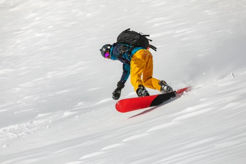 A snowboarder in bright yellow pants and a blue jacket carves through fresh snow on a mountain slope.