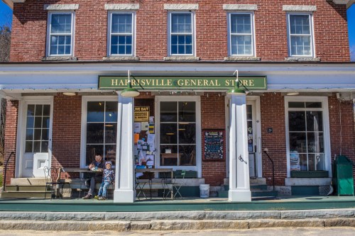 A brick general store with a green sign, featuring a porch where two people sit at a table.